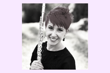 A joyful white woman with vibrant red short hair, wearing a bright smile, holds a flute and gazes directly at the camera.